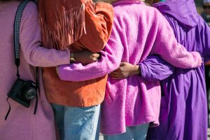 A photo of 4 women with arms around each other's backs - colorful sweaters and scarves