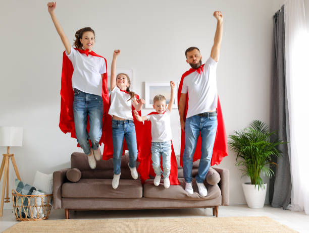 Joyful family in Superhero costumes jumping from sofa
